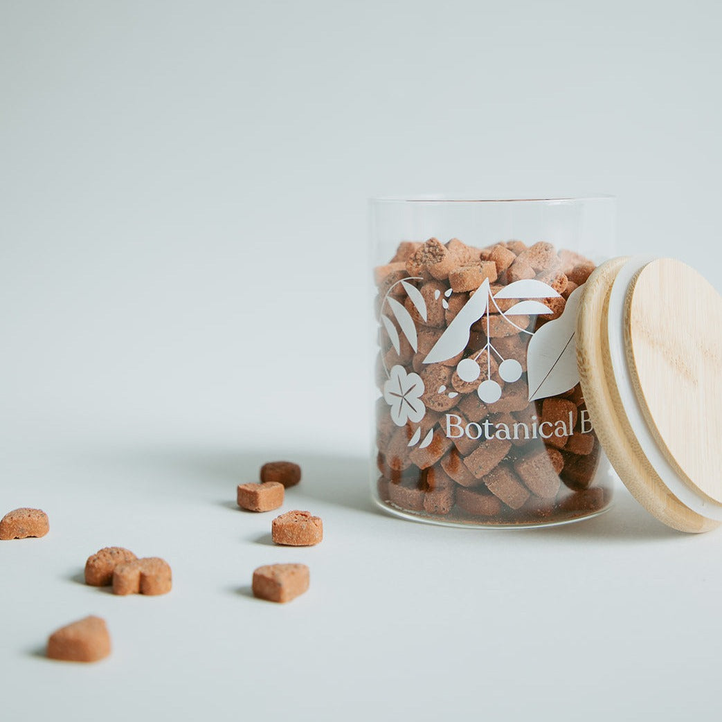 A clear glass jar filled with botanical bones love bites  botanical treat bits, sealed with a wooden lid that has a floral design.